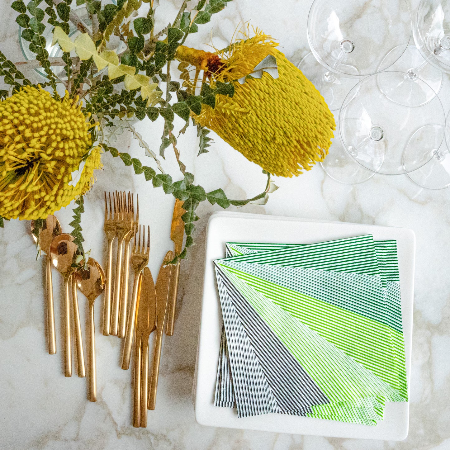 Table setting with gold cutlery, green and white striped napkins, and yellow flowers on a marble surface.