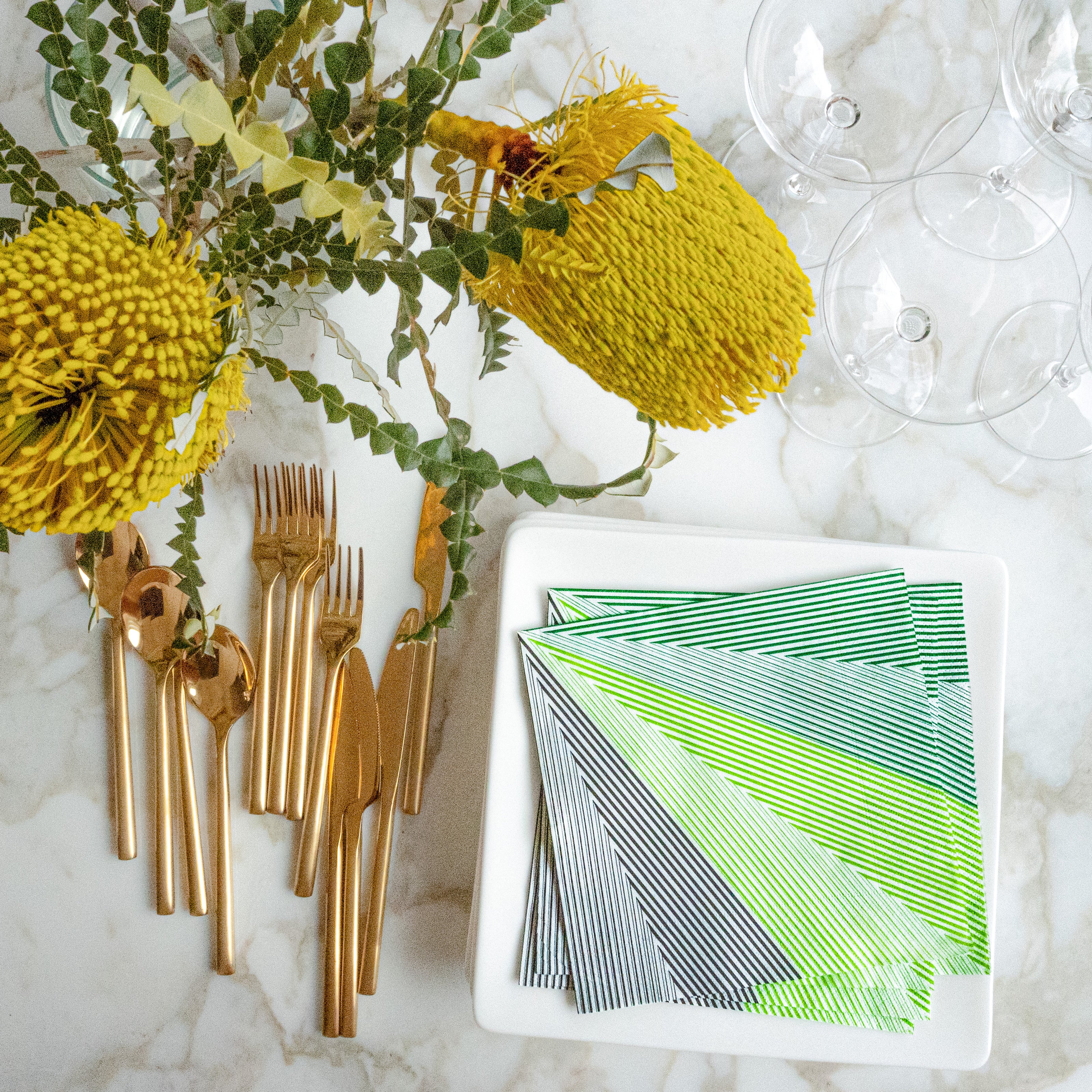 Table setting with gold cutlery, green and white striped napkins, and yellow flowers on a marble surface.